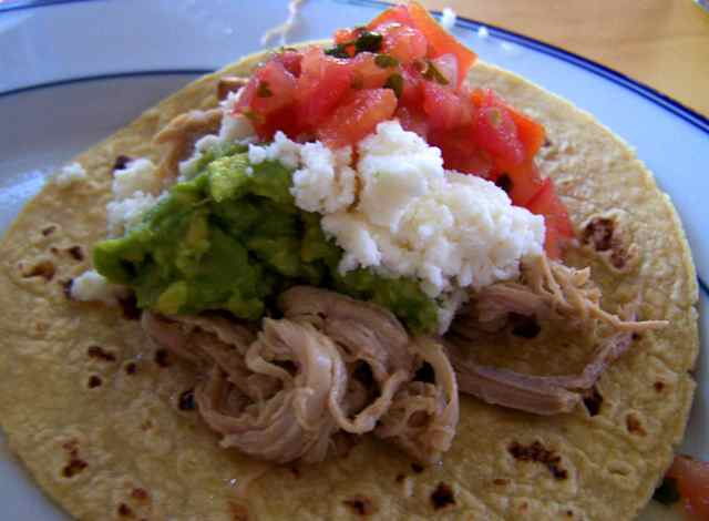 A finished pulled pork taco slow cooker recipe on a tortilla covered in sour cream, guac and salsa