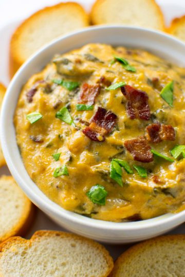 A close up of a white bowl of Spinach Bacon Dip surrounded by bread
