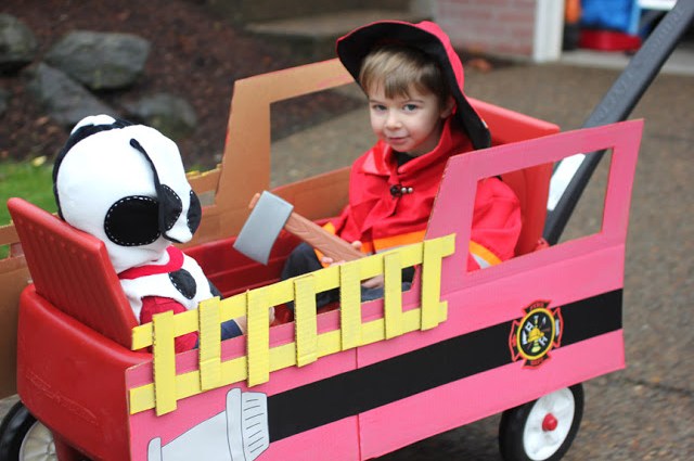 A cardboard box is painting red to look like a fire engine and is worn by a little boy for Halloween