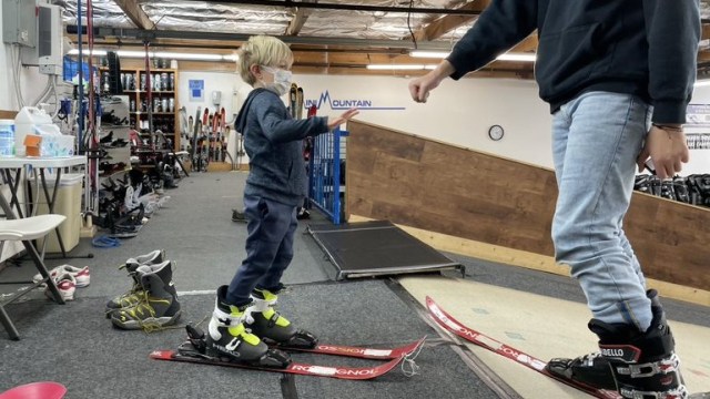 a boy stands at the top of an indoor training ski mountain wearing skis and boots