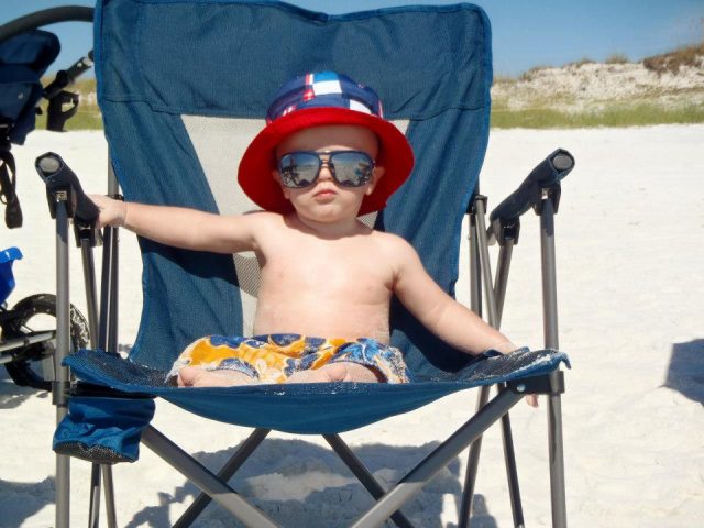 a baby with sunhat and sunglasses sits in a beach chair, using baby beach hacks 