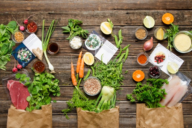 Three paper bags lay on a table with carrots, meat and other ingredients spilling out as part of the Sun Basket meal delivery service