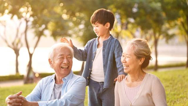 a boy taps his grandfather on the head while grandma sits nearby in a sunny park on Grandparents day with his visiting grandparents