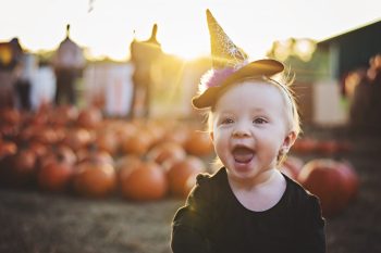 A baby wearing a witches hat sits in front of a pumpkin patch