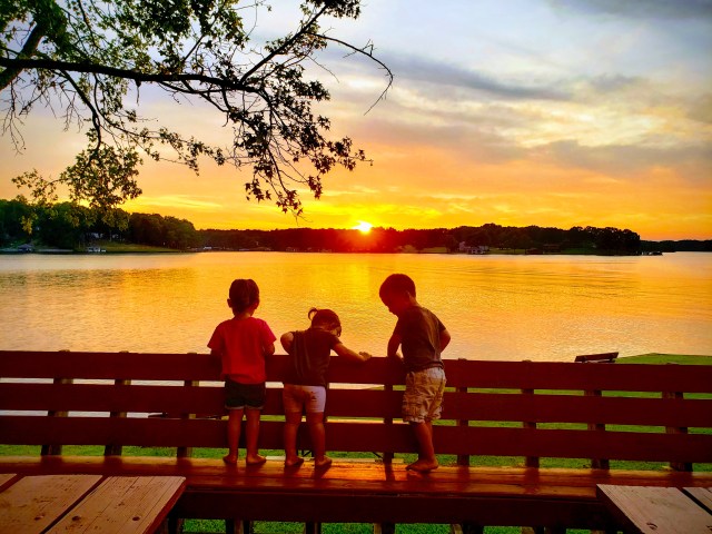 three children standing on bench in front of lake and sunset