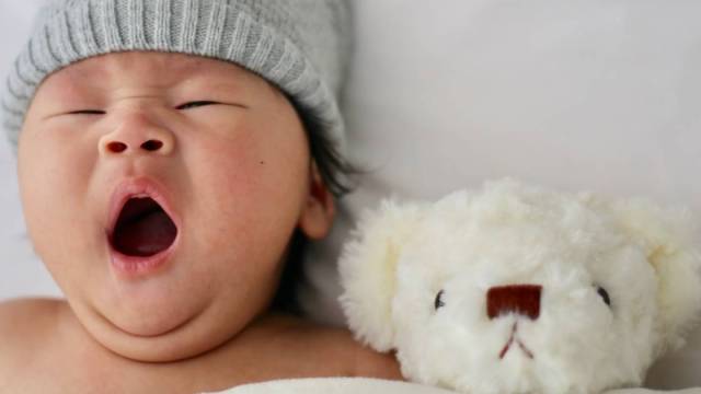 Asian baby yawning in bed next to stuffed animal bear - baby sleep guide