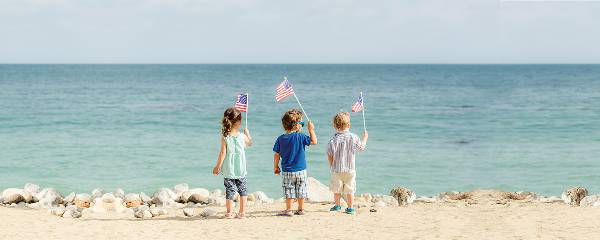 Three kids stand by the beach waving flags on July 4th