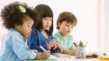 three kids who are kindergarten age draw at a table with markers in the classroom