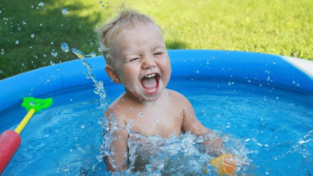 a boy in a baby pool splashing, with toys around him, outdoor water play for babies