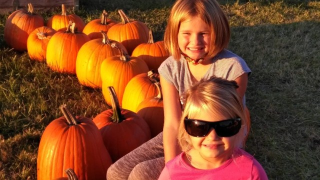 two girls sit in a pumpkin patch squinting against the sun
