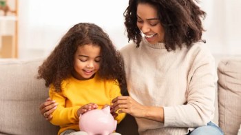 a mom talking to her little daughter who is holding a pink piggy bank