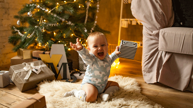 A baby smiles and laughs in front of a Christmas tree as he holds up a present