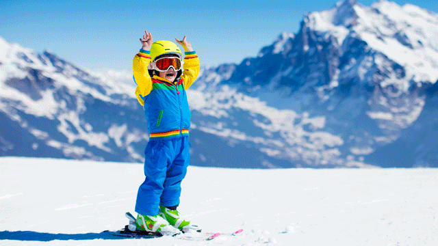 A kid on skis throws his arms in the air in joy while on top of a snowy mountain