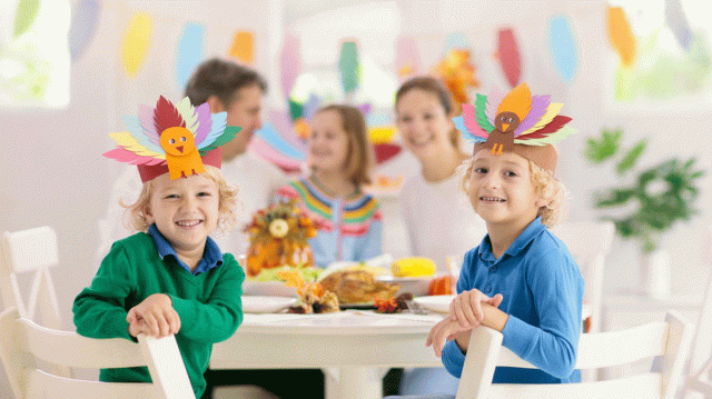Two boys sit at the Thanksgiving dinner table with their family showing off the paper turkey hats they just made