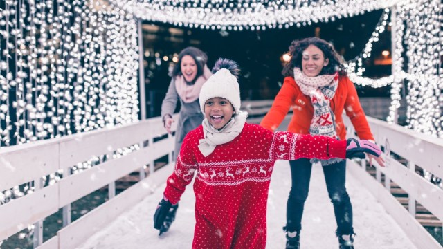 two moms and a child skate around an outdoor ice rink with holiday lights in the background