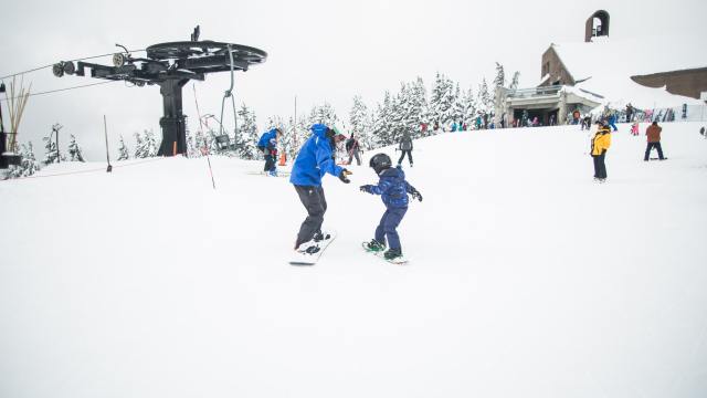 an adult and kid at a ski resort near portland with snow and a lift in the background are going down a hill side by side