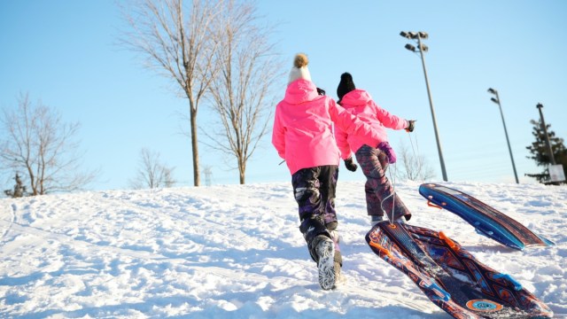 two kids pull sleds up a snowy hill to go sledding in boston with trees and lights in the background