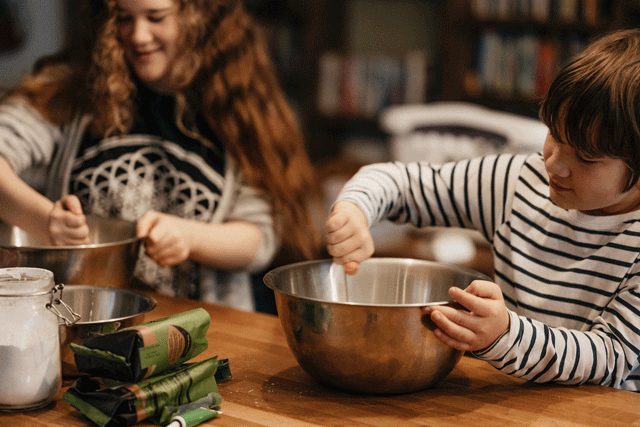 A boy and his friend are in the kitchen stirring ingredients in a silver bowl during a baking themed birthday party