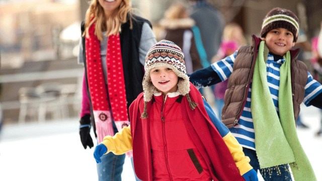 two kids in winter gear are ice skating portland with scarves and hats and a parent in the background