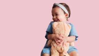 a girl hugs a light brown teddy bear in front of a pink background from a toy store in Boston