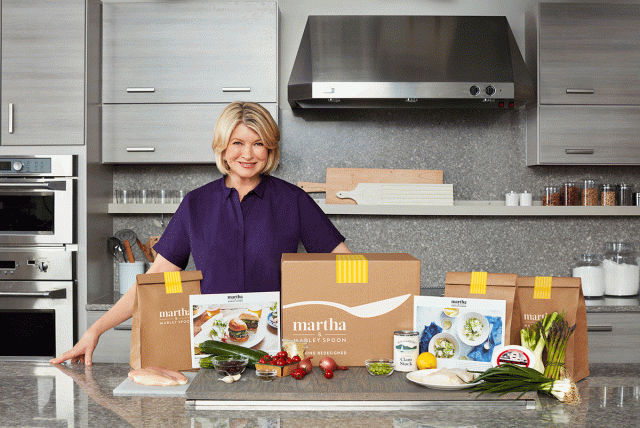 Martha Stewart stands in the kitchen behind a counter filled with boxes and bags of ingredients for her meal delivery service Martha Stewart & Marley Spoon