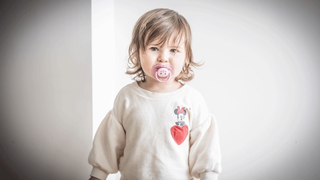 a toddler with a pacifier in her mouth wearing a strawberry shirt is wondering when to get rid of the pacifier