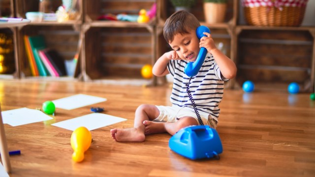 a toddler talks on a blue play phone surrounded by other toys on a wood floor, a language development game