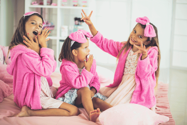 Three little girls in pink bathrobes sit on a bed showing off their nails at a spa birthday party