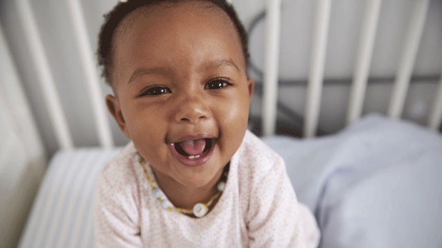 a baby with a big smile sits in a crib waiting for a baby teething biscuit