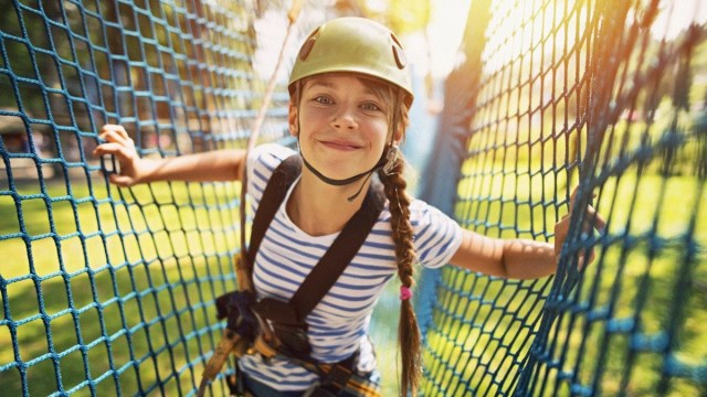 a girl with a helmet and harness smiles as she makes her way through netting in a zipline