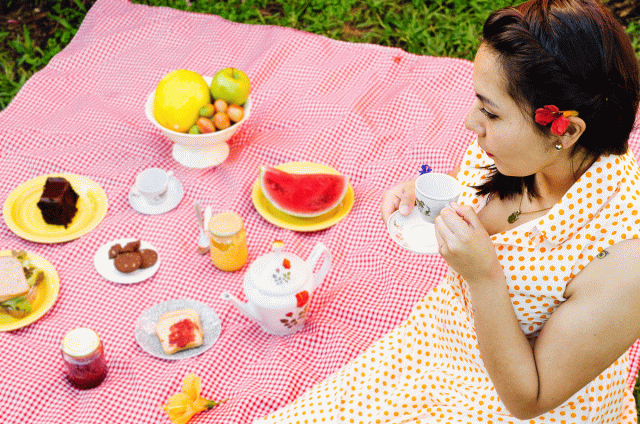 A mother sits on a blanket drinking tea at her daughter's picnic