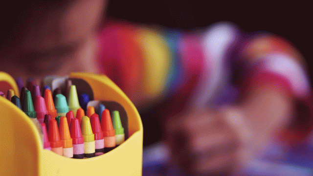 a box of crayons with a kid coloring in the background