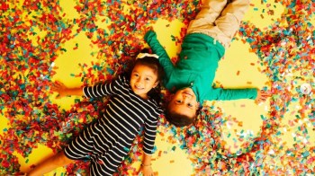 Kids lay on a yellow background with colored confetti all around at Color Factory in NYC, one of the best immersive experiences and pop up museums in the city