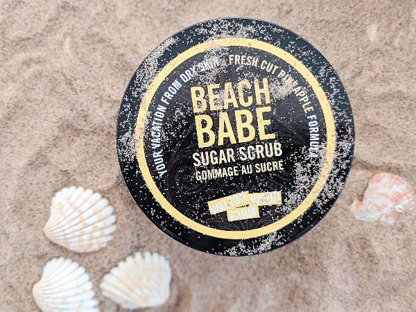 A container of Beach Babe sugar scrub on a sandy beach with shells