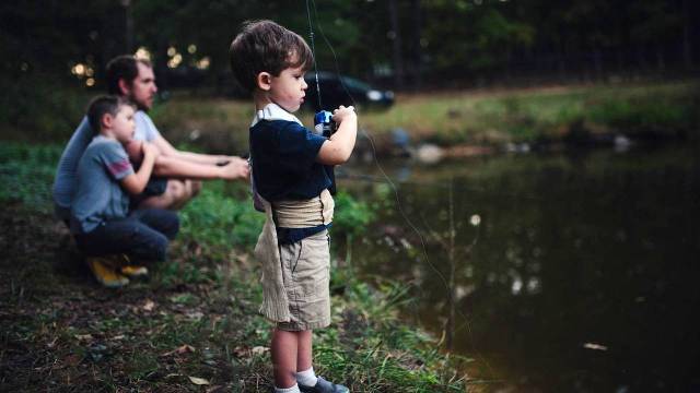 a little boy stands at the edge of a kid-friendly fishing spot with a rod, his parent is in the background
