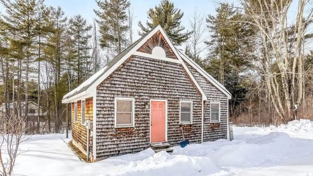A cabin with a red door and a snow shovel popped against it with snow on the ground