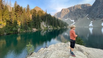 a boy stands on a rock overlooking a lake after a hike in the mountains