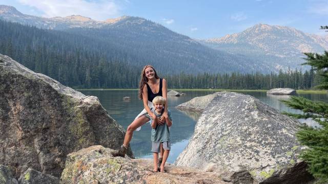 a mom and kid stand on rocks by a lake with mountains in the background and a forest after a lake hike