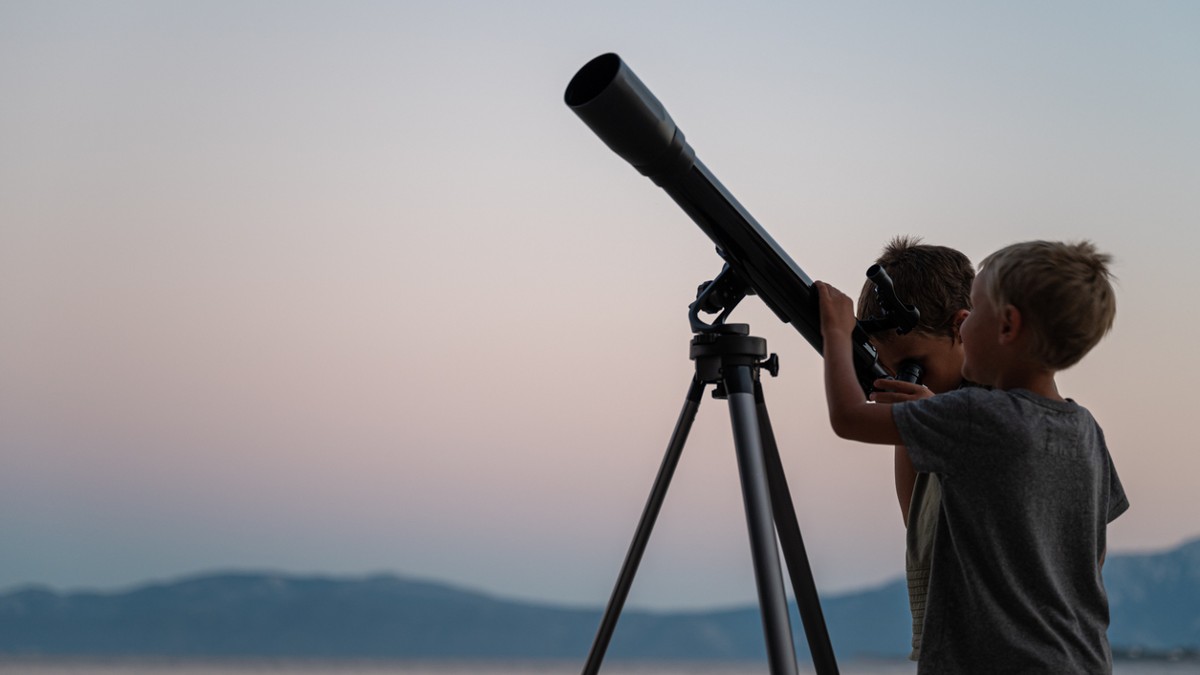 two boys go stargazing looking through a telescope with a light night sky behind them