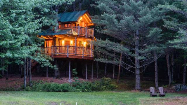 a two story treehouse rentals in new england is lit up at night by the lake with two chairs