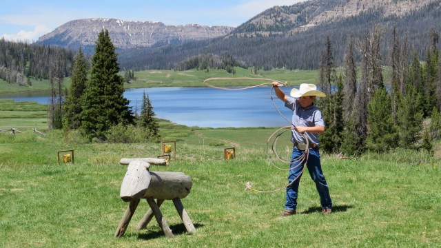 Young boy wearing hat with lasso in a green meadow with lake and mountains behind.