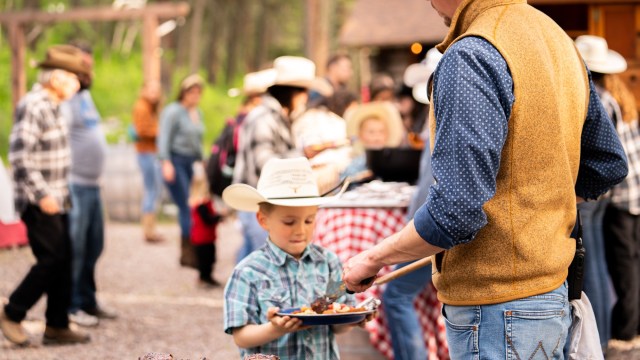 Young boy in plaid shirt and cowboy hat holding a plate of steak while being served by an adult.