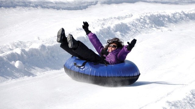 Little girl wearing sunnies, purple jacket and black pants, shoes and gloves, on a blue and black tube, enjoying snowtunign down an oath of snow, with hand and legs in the air.