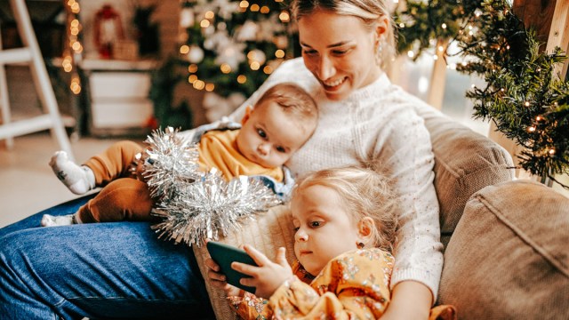 A mom sitting on the couch around the holidays holding her baby with her toddler daughter lying on her lap looking at her phone