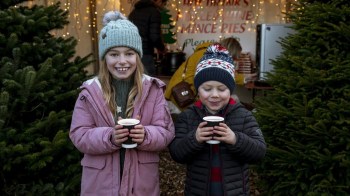 two kids with hot cocoa mugs in front on Christmas trees on Christmas day