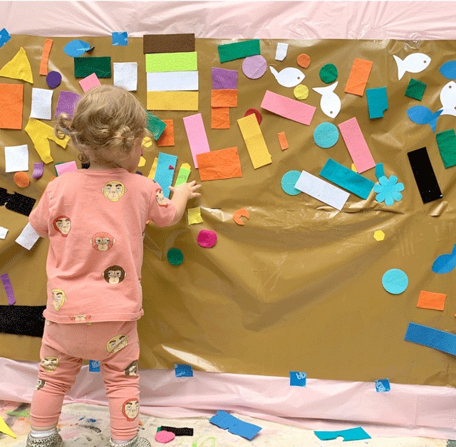 art activities for toddlers can be simple like this sticky paper wall where a kid is hanging paper on a brown felt board
