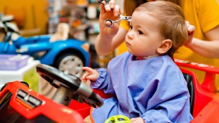 a child with a blue apron on sits in a car chair during baby's first haircut