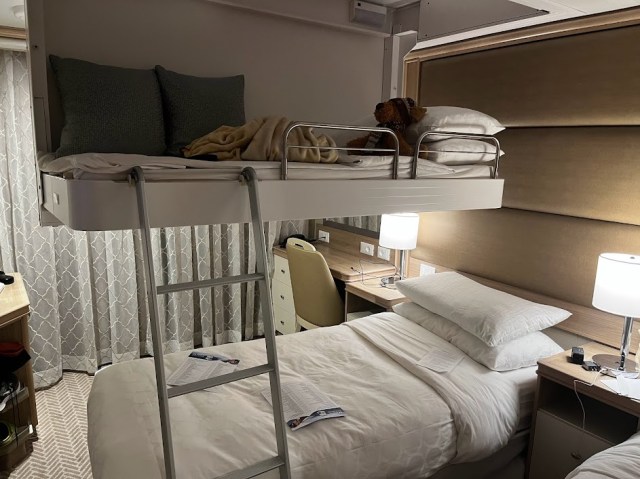 Balcony suite on Sky Princess cruise ship. Two twin beds with one additional twin bed lowered from ceiling with ladder attached. Night stands between beds have lights turned on.