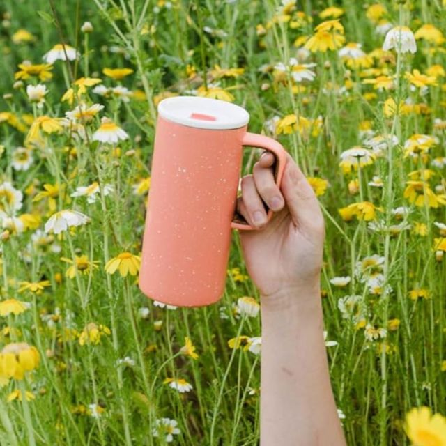 arm holding a peach camp travel mug in front of a field of yellow flowers