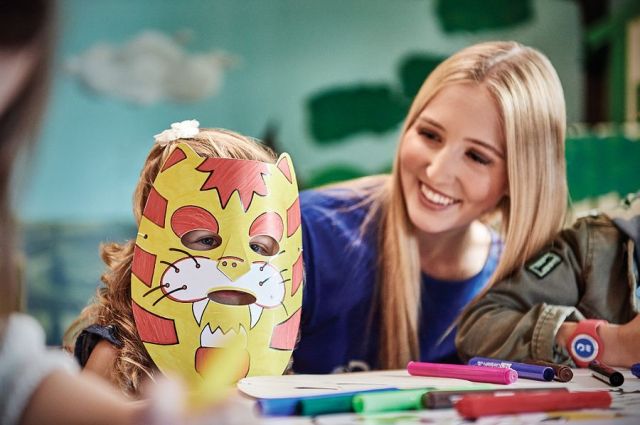 Blond woman with blue shirt sitting at table and smiling at small child with tiger paper mask on face. Table has craft supplies laid out in front of them.